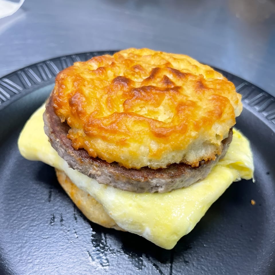A breakfast sandwich with a golden biscuit, sausage patty, and melted cheese on a black plate.