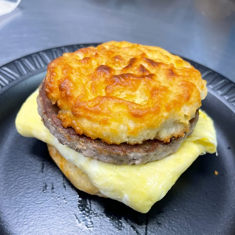 A breakfast sandwich with a golden biscuit, sausage patty, and melted cheese on a black plate.